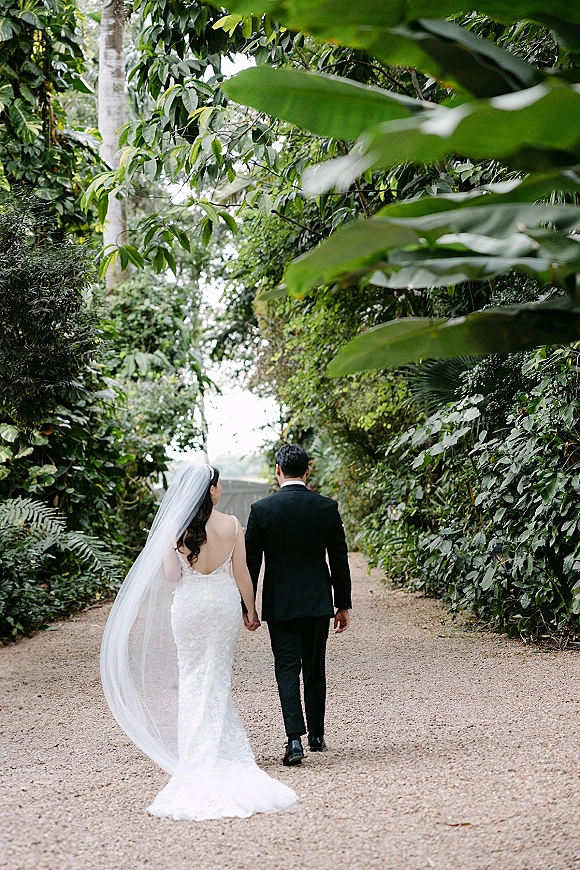 Wedding couple portrait of bride and groom walking away holding hands, her long veil and lace dress trailing on a tree-lined garden path