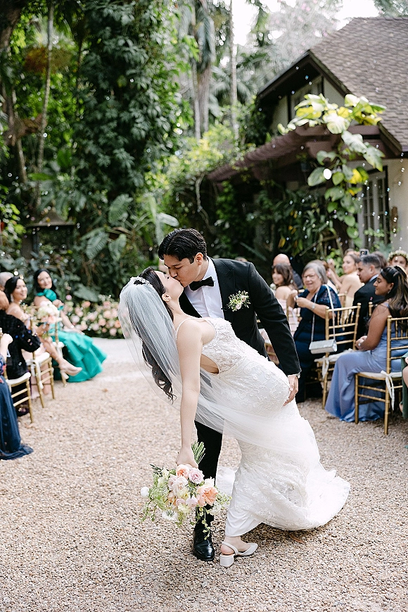 Wedding kiss portrait of groom dipping bride in lace dress and veil, bouquet in hand, as guests watch along a garden aisle