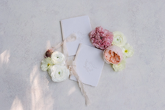Invitation suite flat lay with deckled edge paper tied in chiffon ribbon on a light stone surface, styled with ranunculus blooms