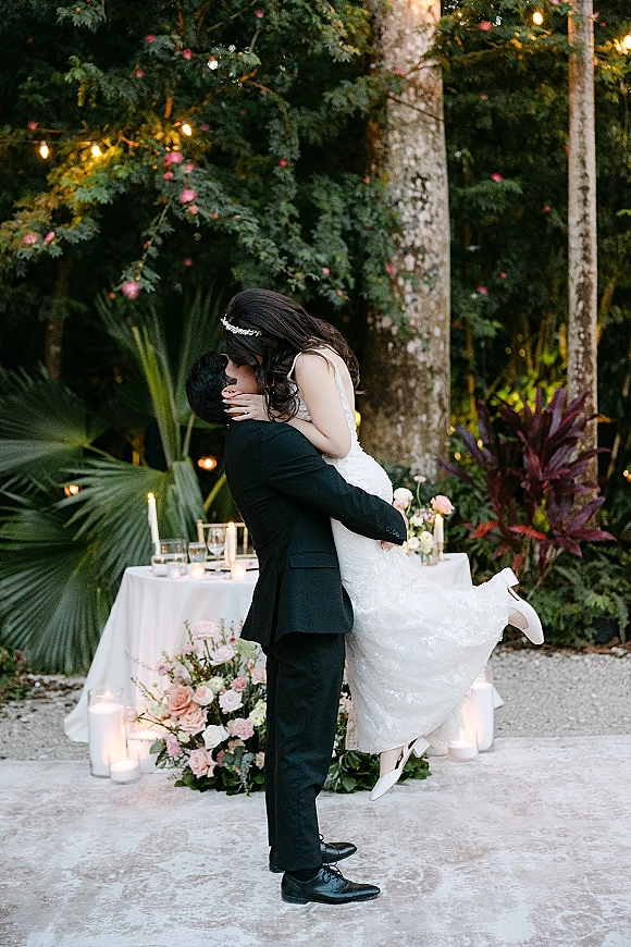 Wedding kiss portrait of groom lifting bride for a dip kiss in a garden, her strapless lace dress and ring visible amid greenery