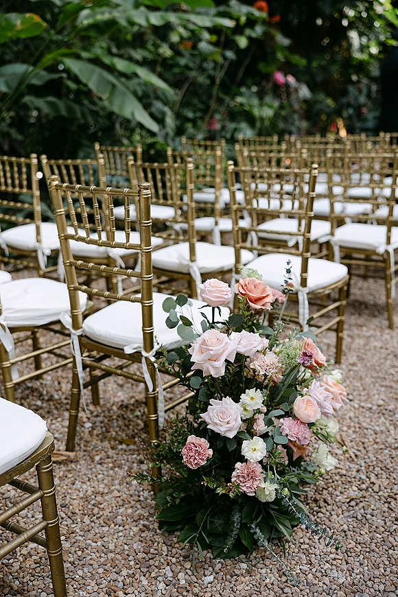 Ceremony seating with gold chiavari chairs and white cushions, tied with white ribbons beside aisle florals with blush roses and eucalyptus in a garden setting