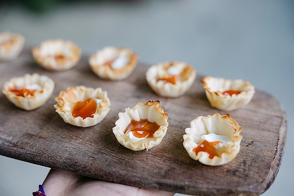 Wedding appetizers on a wooden serving board, mini tart shells with creamy filling and orange jam, held on a tray against a neutral wall