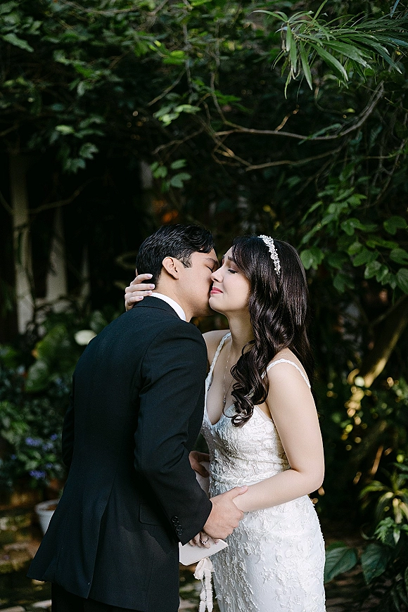 Wedding kiss as bride in a lace gown cradles the groom’s face while he holds her waist, framed by lush garden greenery