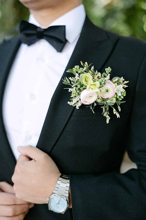 Groom boutonniere with ranunculus pinned to a classic black tuxedo lapel, bow tie and wristwatch visible against greenery outdoors