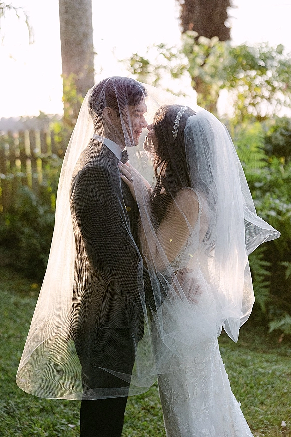 Wedding couple portrait with veil draped over the couple as they embrace forehead to forehead, backlit at sunset in a garden lawn near trees