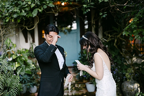 First look moment as bride in a lace wedding dress reads a letter to groom in black suit by a cottage blue door and garden greenery