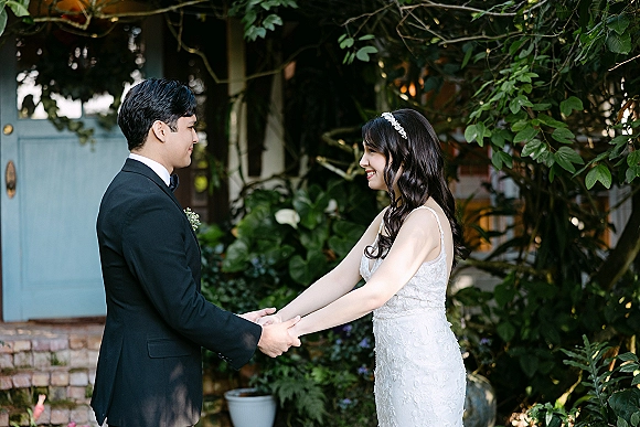 First look moment as bride and groom holding hands by a blue door on brick steps, garden greenery behind, her lace dress and headband visible