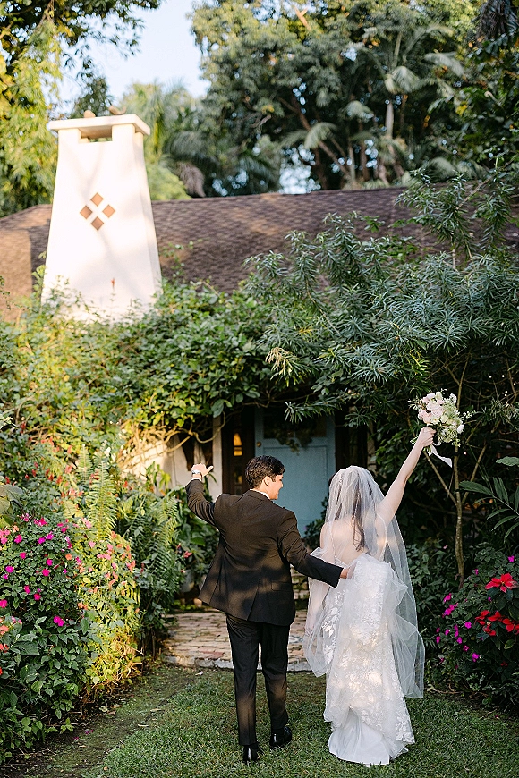 Wedding exit as newlyweds walk away hand in hand down a garden path, bride raising her bouquet with veil trailing past a cottage backdrop