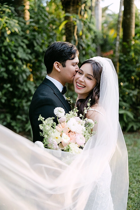 Couple portrait of groom kissing bride’s cheek as she laughs, holding a bouquet with a flowing bridal veil in a green garden setting
