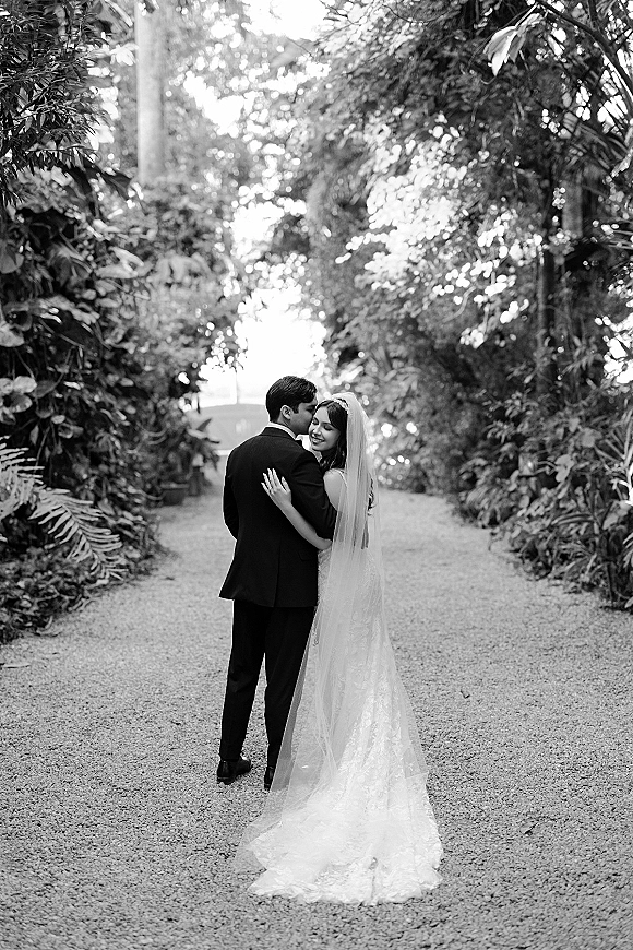 Couple portrait in black and white, groom kissing bride’s forehead as she smiles on a tree-lined garden path with a flowing veil train
