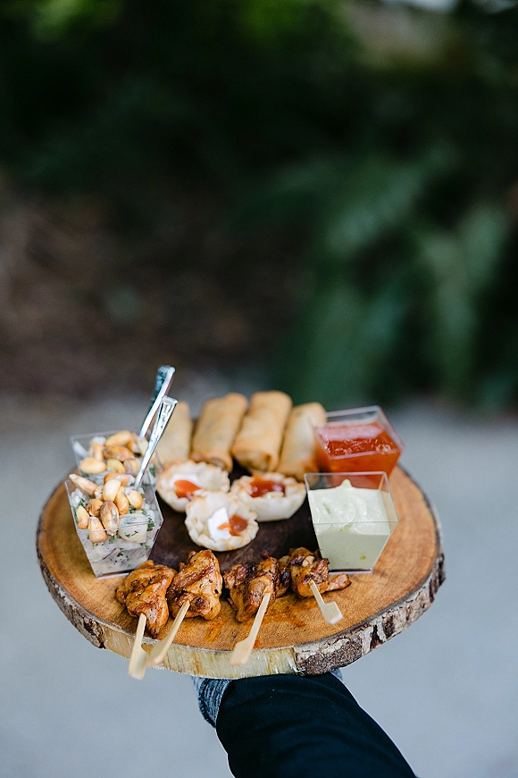 Wedding cocktail appetizers on a wood serving board with skewers, mini spring rolls, and dipping sauce cups against blurred outdoor greenery