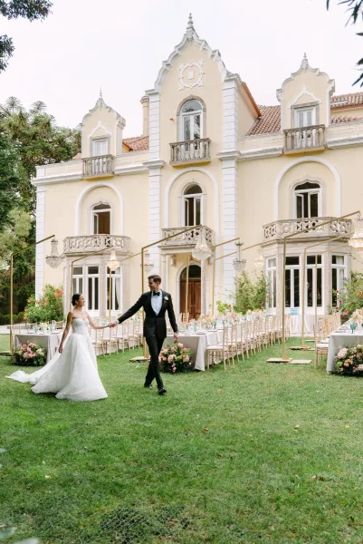 Couple portrait of bride and groom holding hands in a garden lawn, her strapless lace wedding dress and his tuxedo before a historic villa facade