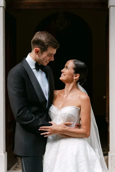 Couple portrait of bride and groom portrait holding hands, her lace dress and veil framed by an arched doorway and stone columns