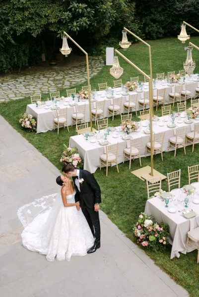 Wedding kiss portrait of bride in strapless gown and lace veil with groom in tuxedo beside outdoor wedding reception tables on a garden patio lawn