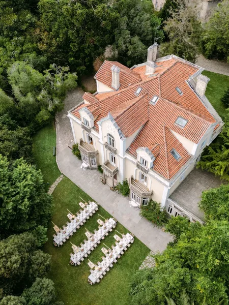 Outdoor reception setup with long banquet tables in white linens, floral centerpieces, and taper candles on a stone patio by a historic villa