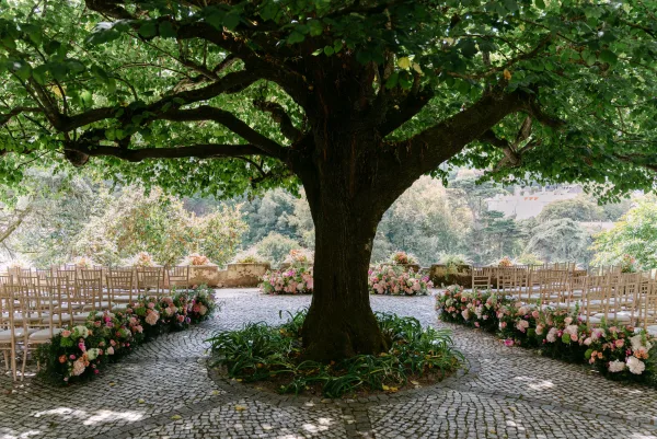 Outdoor ceremony setup under a large tree, wooden chairs in a circle around pink and white floral aisle arrangements on cobblestones