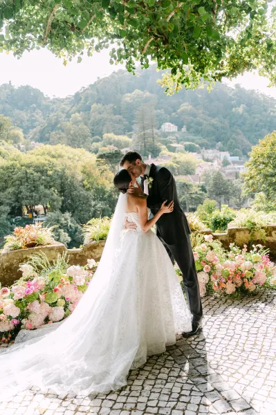 Wedding kiss portrait of bride and groom kissing, her long veil and lace train flowing on a cobblestone terrace with hillside village view
