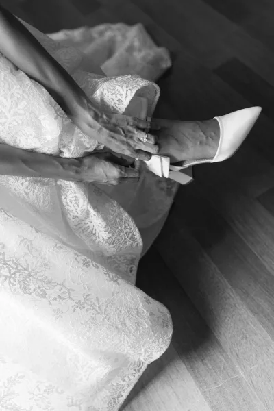 Bridal shoes as bride puts on white pointed toe heels with ankle strap, wedding ring visible beside lace dress on wood floor