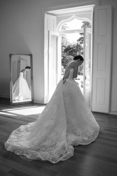 Bridal portrait of a bride by window in a lace open-back gown with long train, drop earrings, and floor mirror in airy white room