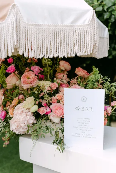 Wedding bar signage with a printed menu beside roses and hydrangea on a white bar cart under a fringe umbrella on the lawn