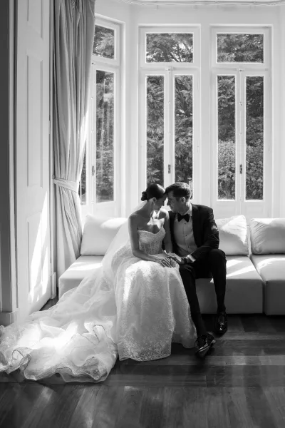 Couple portrait of bride and groom holding hands and touching foreheads on a white sofa, lace gown and veil in window light