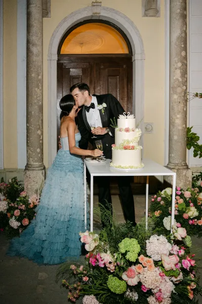 Wedding cake cutting as bride in a strapless ruffled gown and tuxedo groom slice a three tier cake with floral accents in a stone courtyard entrance