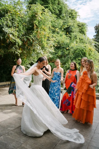 Bride with bridesmaids admiring her strapless lace veil and gown on a stone terrace, colorful dresses framed by garden trees under clouds