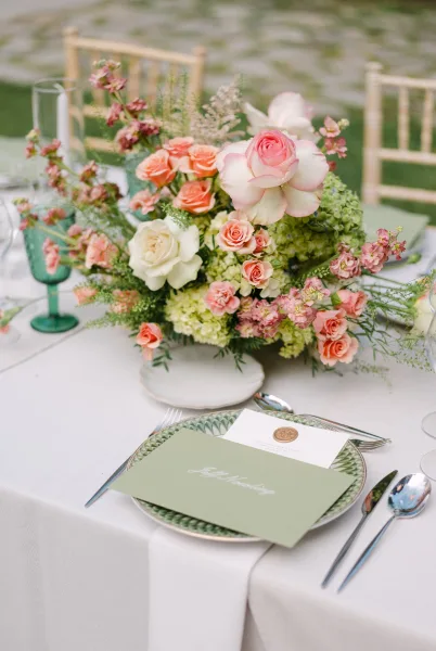 Reception tablescape with wedding floral centerpiece of roses, hydrangea, and greenery, green goblet place setting on an outdoor lawn