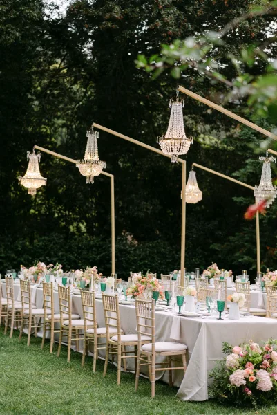 Reception tablescape with crystal chandeliers over long banquet tables, white linens, teal goblets, candles, and flowers on a garden lawn