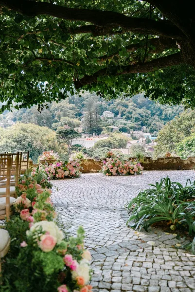 Ceremony aisle design with outdoor wedding aisle flowers bordering cobblestone, wooden chairs beneath a tree canopy in a courtyard