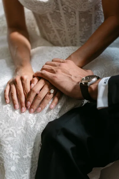 Wedding hands with bride and groom holding hands, featuring an oval diamond engagement ring and lace dress detail in soft natural light