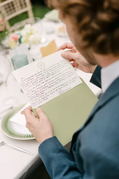 Wedding letter and handwritten wedding note resting on an envelope beside a patterned dinner plate, fork, and glassware at a reception table on the lawn