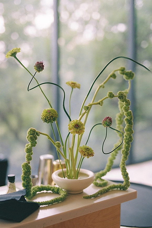Floral centerpiece of monochromatic green blooms in a vase on a table with napkin and metal cups by a window with greenery outside