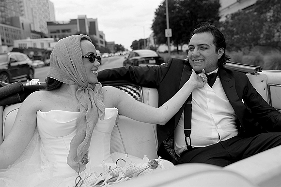 Couple portrait of newlyweds in convertible, bride in strapless dress and sunglasses touching groom’s face on a city street backdrop