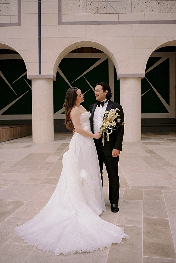 Couple portrait of bride and groom embracing, bride holding a white bouquet in a strapless gown under stone arches in a tiled courtyard