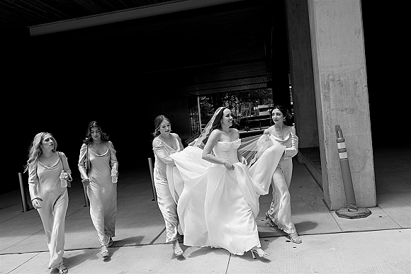 Bride with bridesmaids walking together as bridesmaids helping bride adjust her veil and hold her dress on a city walkway by columns