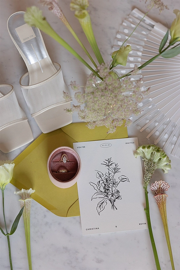 Invitation suite wedding invitation flatlay arranged with a mustard envelope, rings in a velvet box, bridal heels, and flowers on marble surface