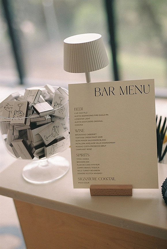 Bar menu sign and wedding bar menu card in a wooden holder beside a table lamp and matchboxes, with cocktail illustrations by a window
