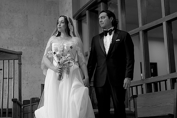 Couple portrait in black and white wedding portrait style, bride in lace veil with bouquet holding hands with groom in tuxedo on stairs