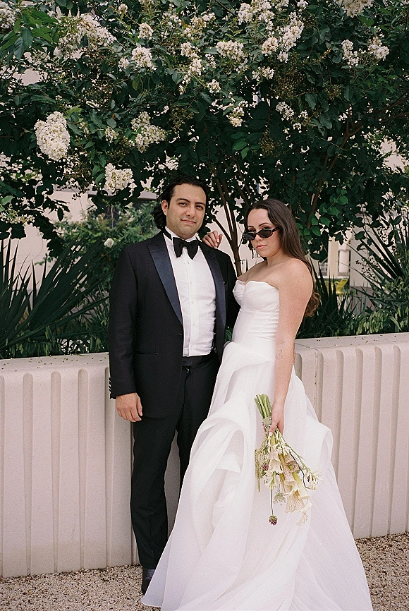 Couple portrait of bride in a strapless wedding dress holding a hand-tied bouquet beside groom in black tuxedo with sunglasses under a flowering tree