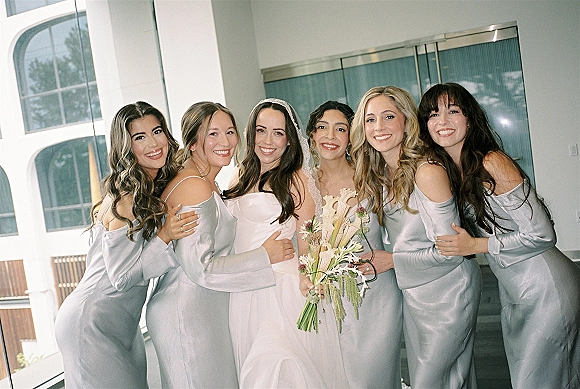 Bridesmaid group photo of bride with bridesmaids in satin bridesmaid dresses, holding bouquets in a modern indoor venue by large windows