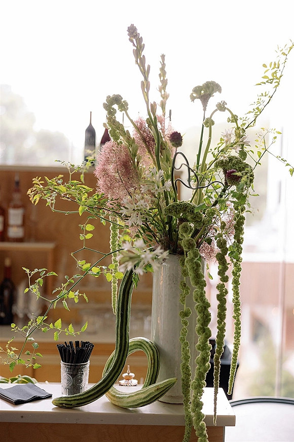 Wedding centerpiece in a tall vase with wildflowers and hanging amaranthus on a reception bar, with glassware, bottles, and window light