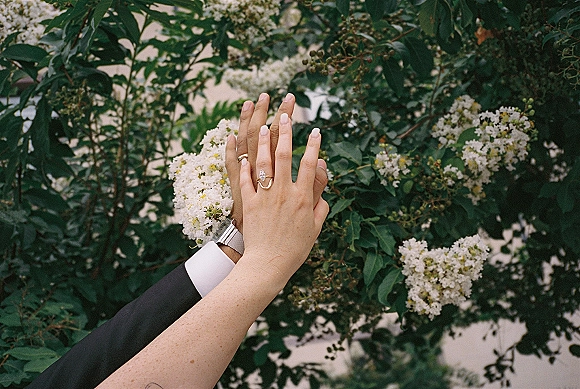 Wedding rings with engagement ring close up on manicured hands, groom’s wristwatch and suit cuff against green foliage and white blooms