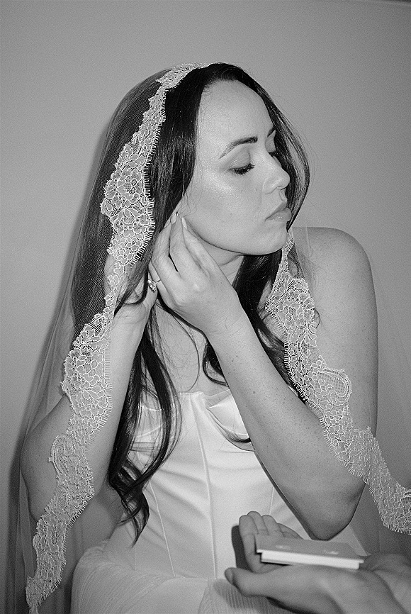 Bridal portrait in black and white bridal photo style of a bride in side profile putting on earrings, lace veil framing her face indoors