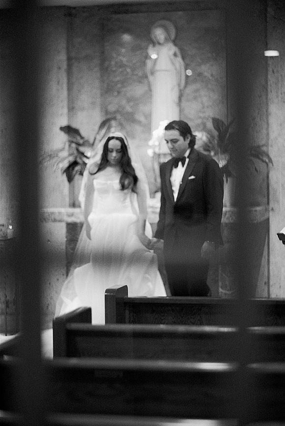 Wedding recessional as bride and groom walk the aisle hand in hand, her long veil trailing past church pews toward the altar statue