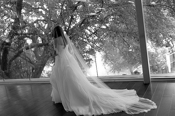 Bridal portrait of a bride with long veil holding a bouquet, gazing out floor-to-ceiling windows with trees beyond and columns behind