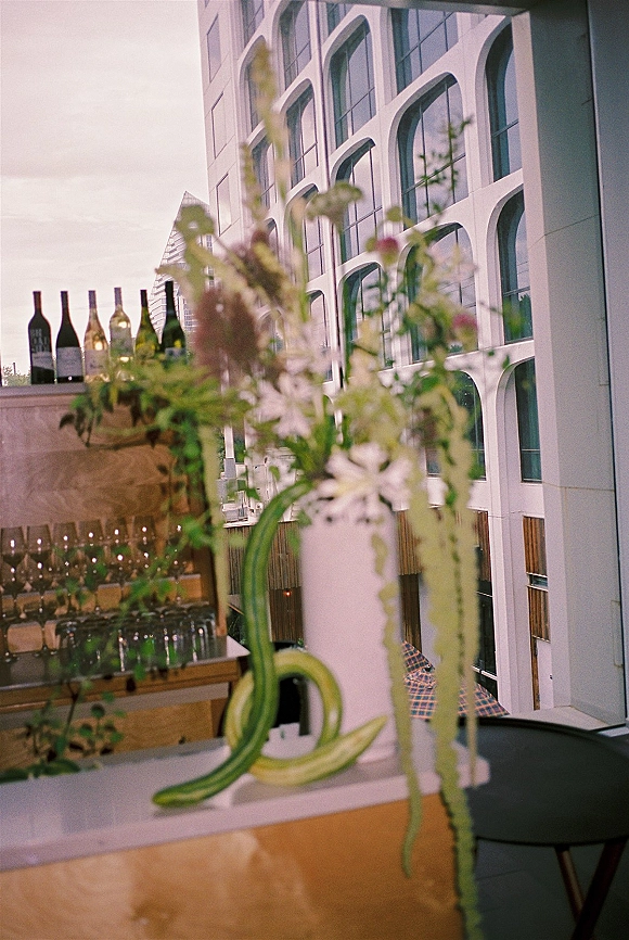 Wedding bar setup with a wedding cocktail bar display of wine bottles and stemware on shelves, plus a tall white vase with greenery on a rooftop terrace