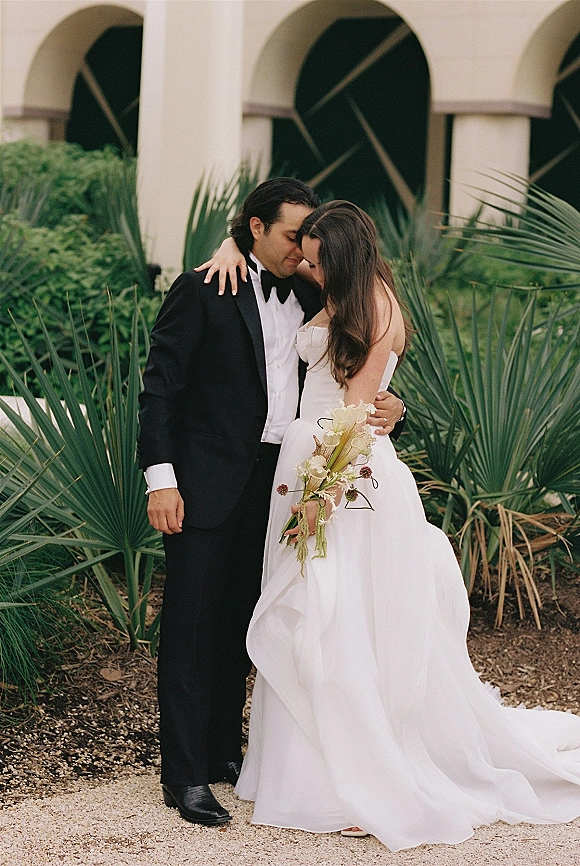 Couple portrait of bride and groom embrace with forehead touch, her strapless gown and calla lily bouquet on a palm-lined path by arches