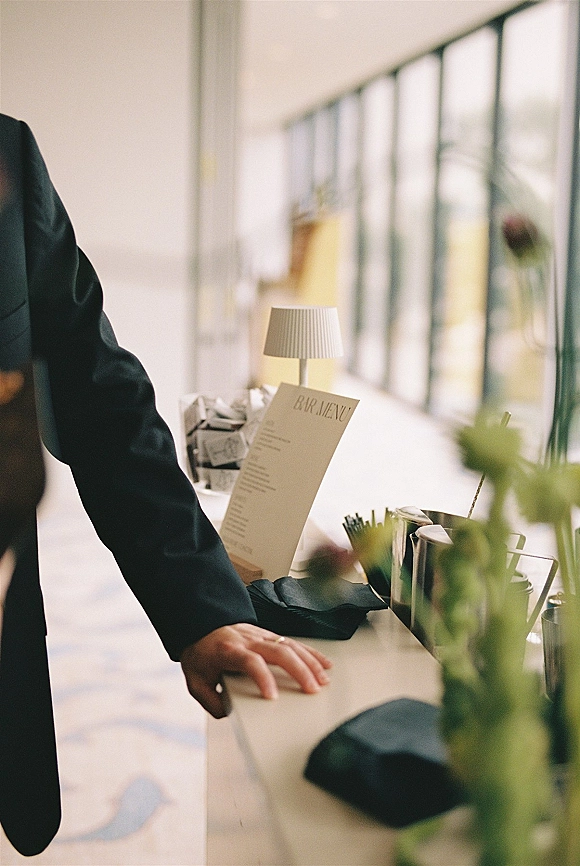 Wedding bar setup with a wedding bar menu, bar tools, ice bucket and glasses on an indoor counter by large windows in daylight