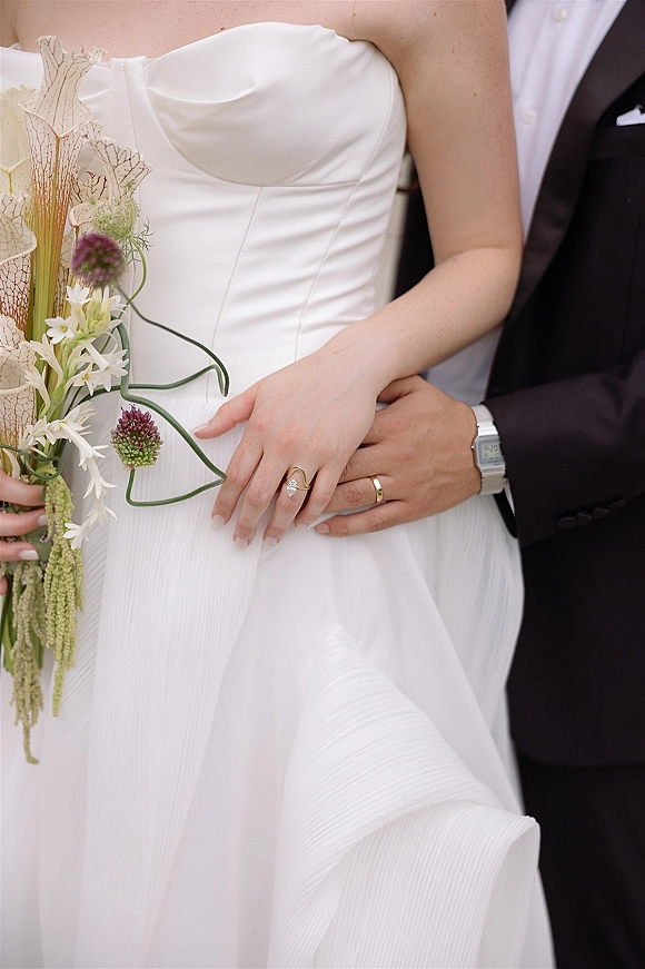 Wedding couple portrait of bride and groom embrace, his hand on her waist showing rings, strapless gown and bouquet on neutral backdrop